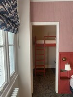A room with a bunk bed, a window with a patterned curtain, a radiator, and a wall-mounted lamp.