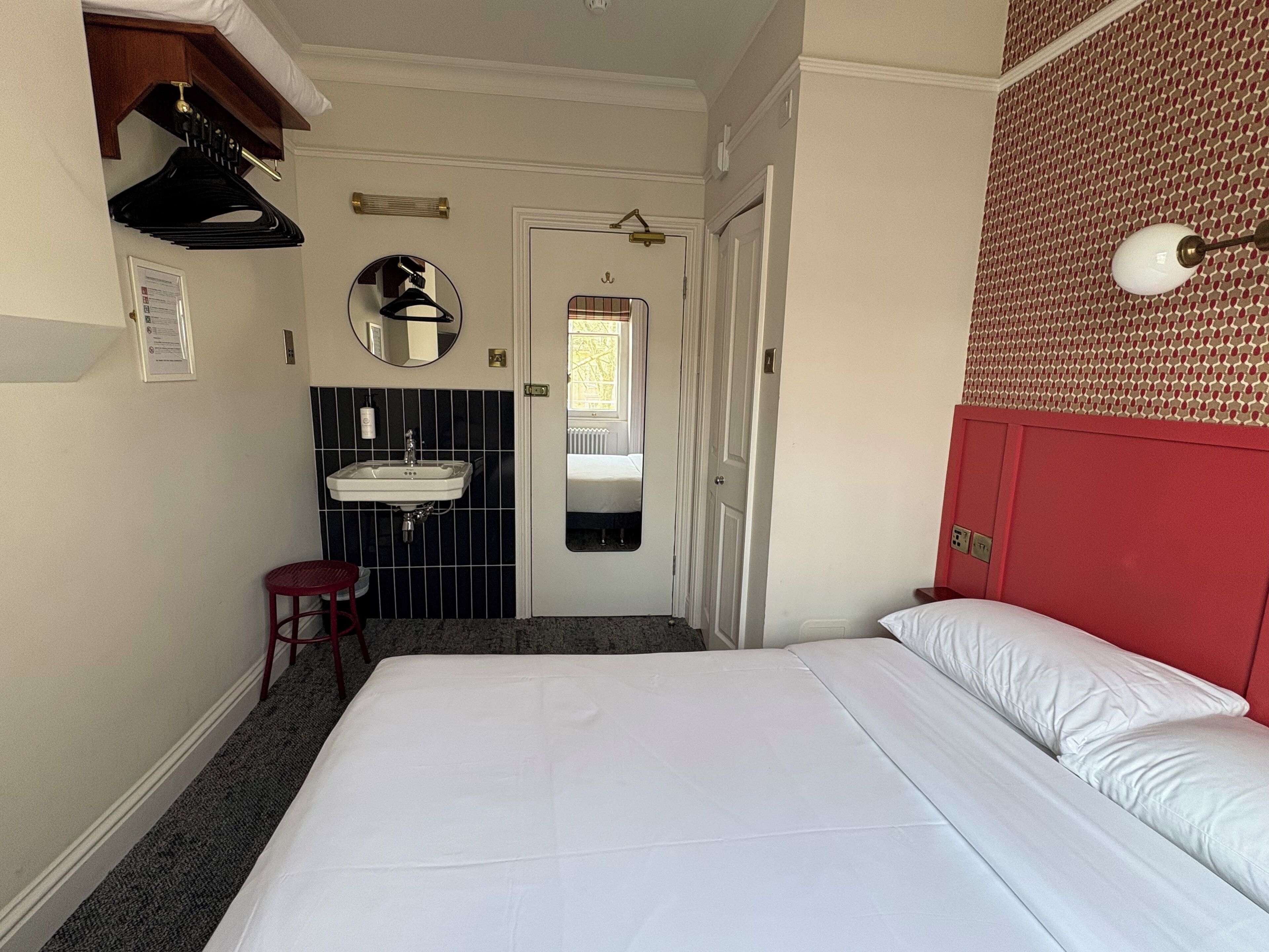 A hotel room with a bed, a red headboard, a white wall, a black and white tiled bathroom, and a red stool.