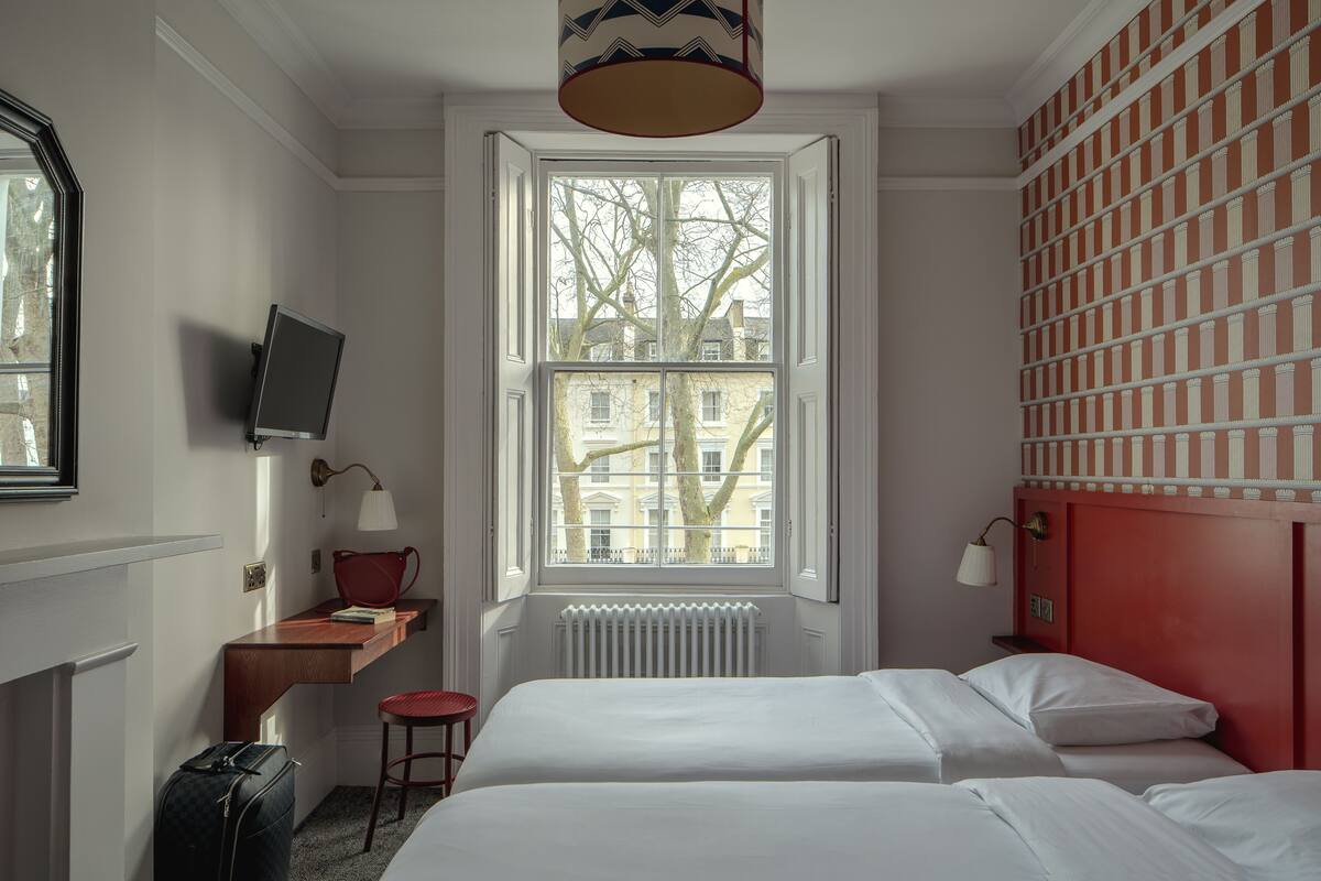 A hotel room with a bed, a desk with a chair, a television, and a window with a view of buildings and trees.