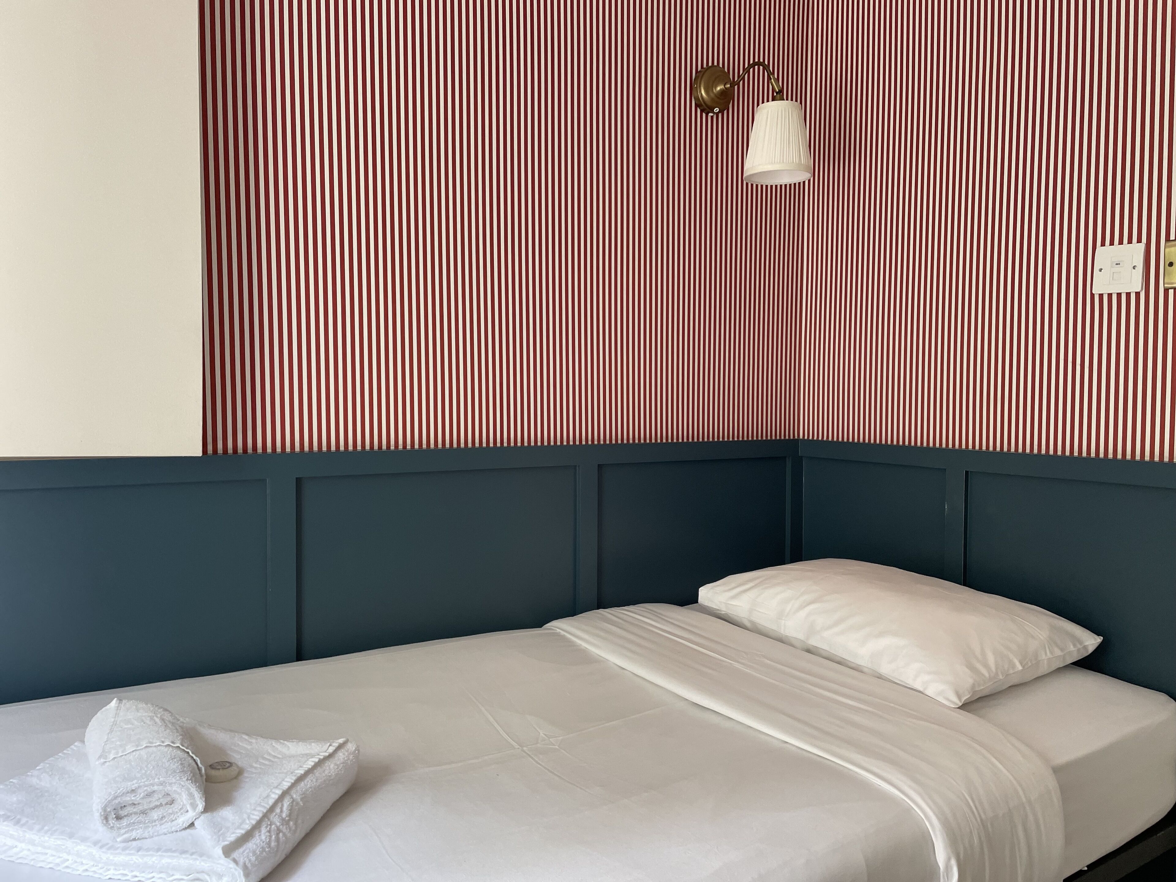 Ein ordentlich bezogenes Bett mit weißen Leinen und einem aufgerollten Handtuch, eine an der Wand befestigte Leuchte und ein gestreiftes Tapetenmuster.