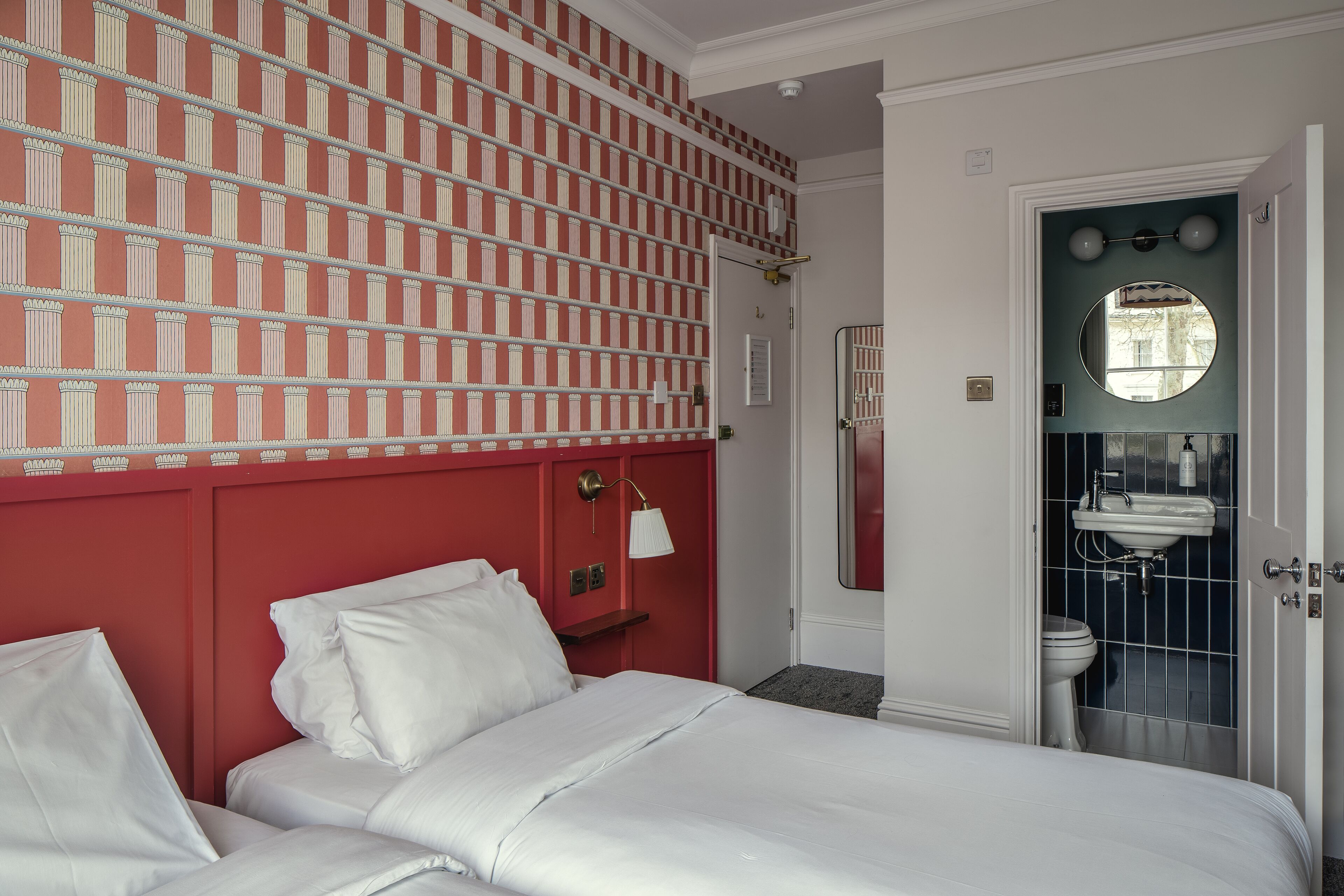 A hotel room with a red headboard, white bedding, a mirror above the sink, and a door leading to a bathroom.
