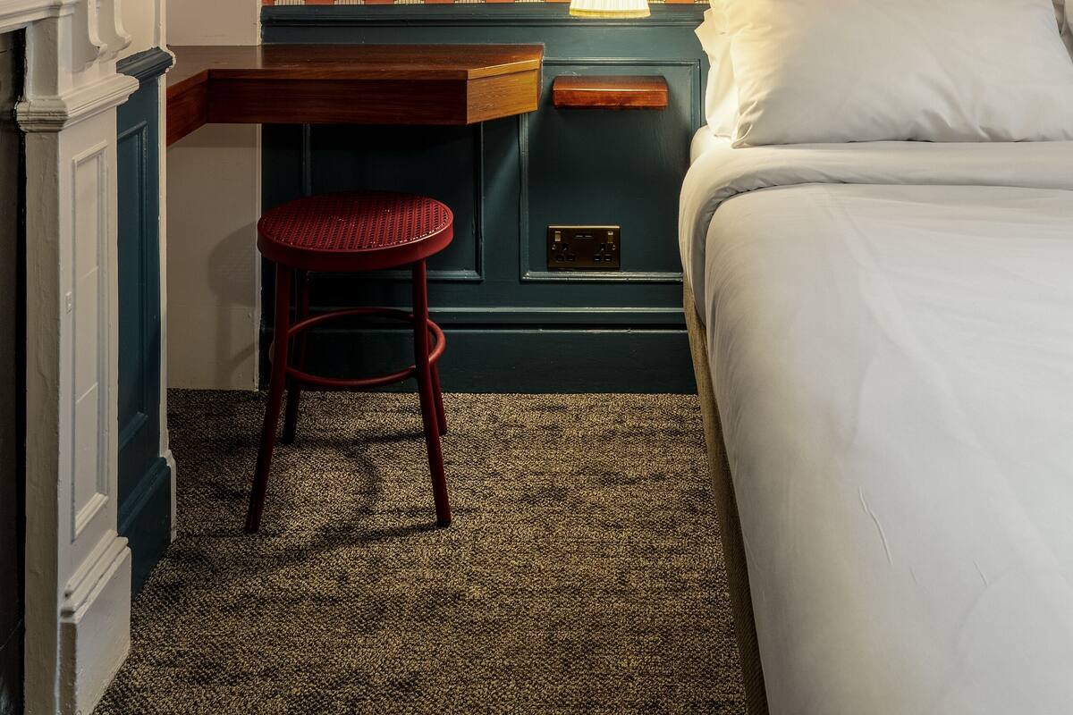 A hotel room with a patterned wallpaper, a bed, a desk with a chair, and a nightstand with a lamp.