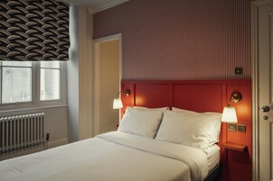 Ein Hotelzimmer mit einem Bett, zwei Nachttischlampen, einem Heizkörper, einem Fenster mit einem gemusterten Rollo und einem roten Kopfteil.