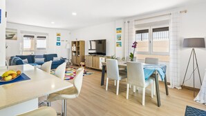 A modern living room with a sofa, dining table, and chairs. There is a TV, a cabinet, and a window with curtains.