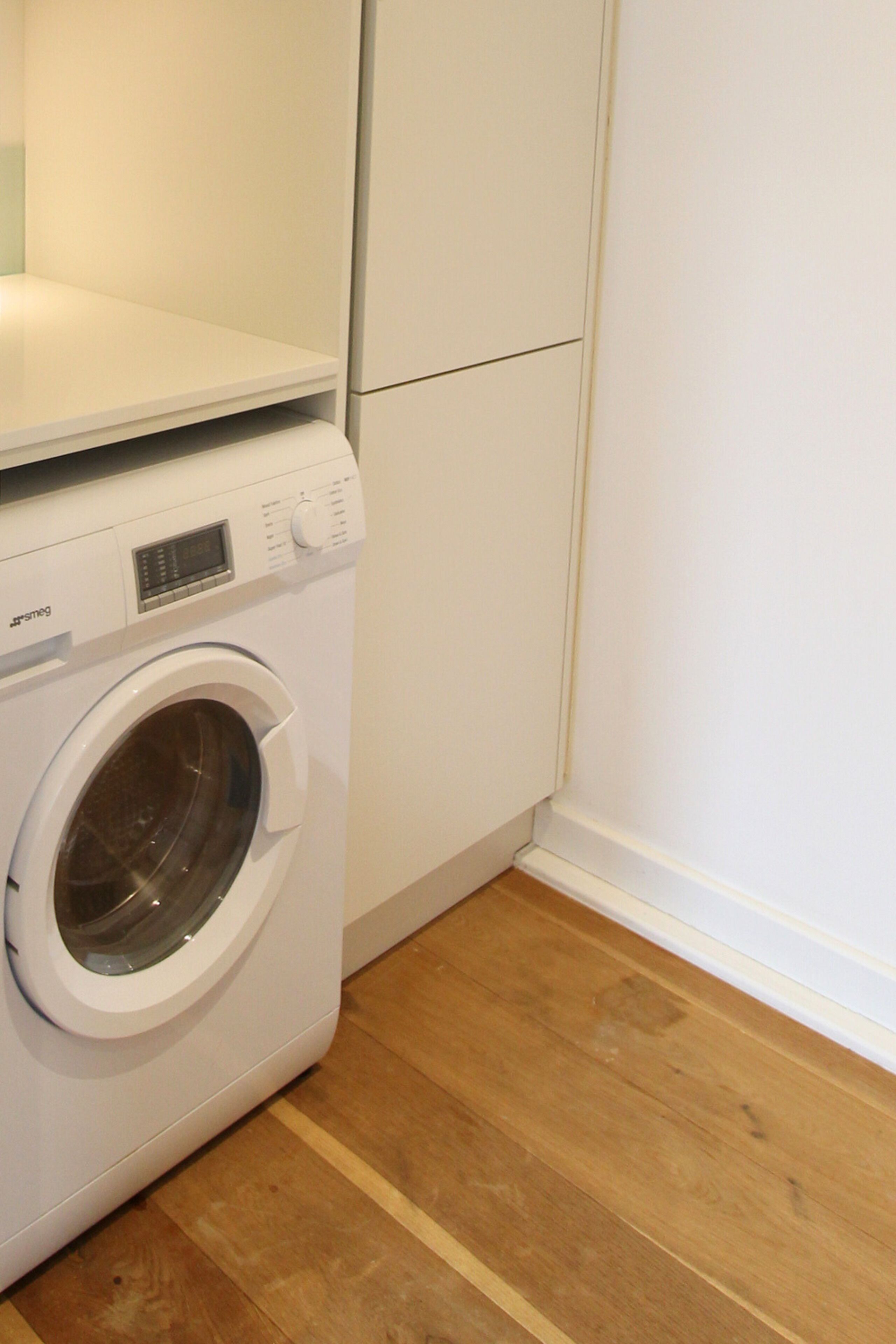 A front view of a washing machine in a corner of a room with wooden flooring and white walls.