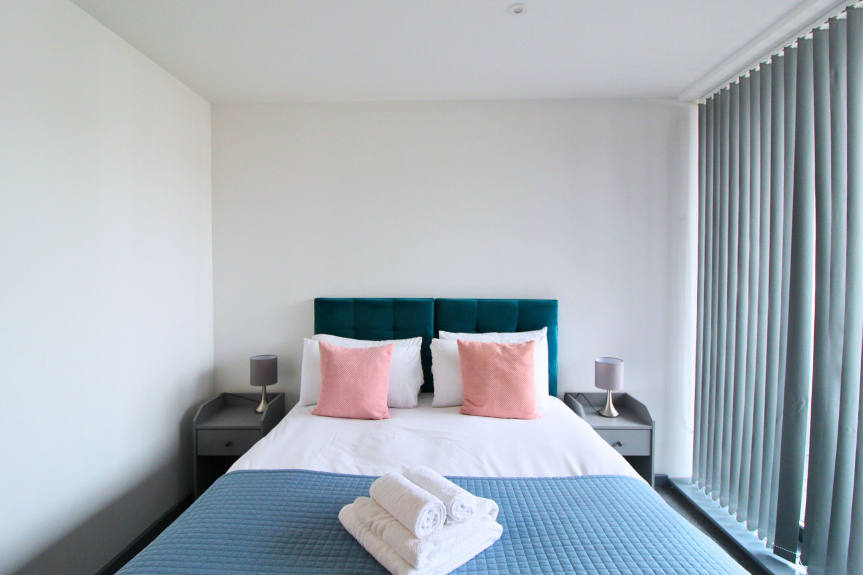 A neatly made bed with a blue quilt, two pink pillows, and a teal headboard. There are two bedside tables with lamps and a window with blinds.