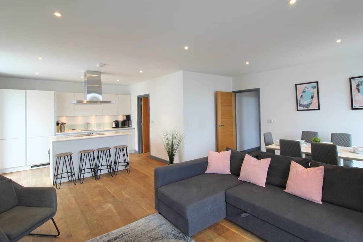 A modern living room with a grey sofa, a dining area with a table and chairs, and a kitchen with white cabinets and a bar stool area.