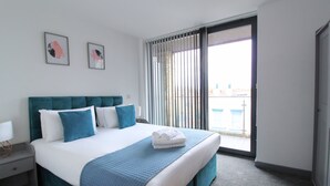 A hotel room with a large bed, a nightstand, a window with blinds, and two framed pictures on the wall. - Canning Town by Viridian Apartments (London)