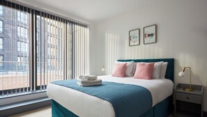 A modern hotel room with a large bed, a view through floor-to-ceiling windows, and a nightstand with a lamp. - Canning Town by Viridian Apartments (London)