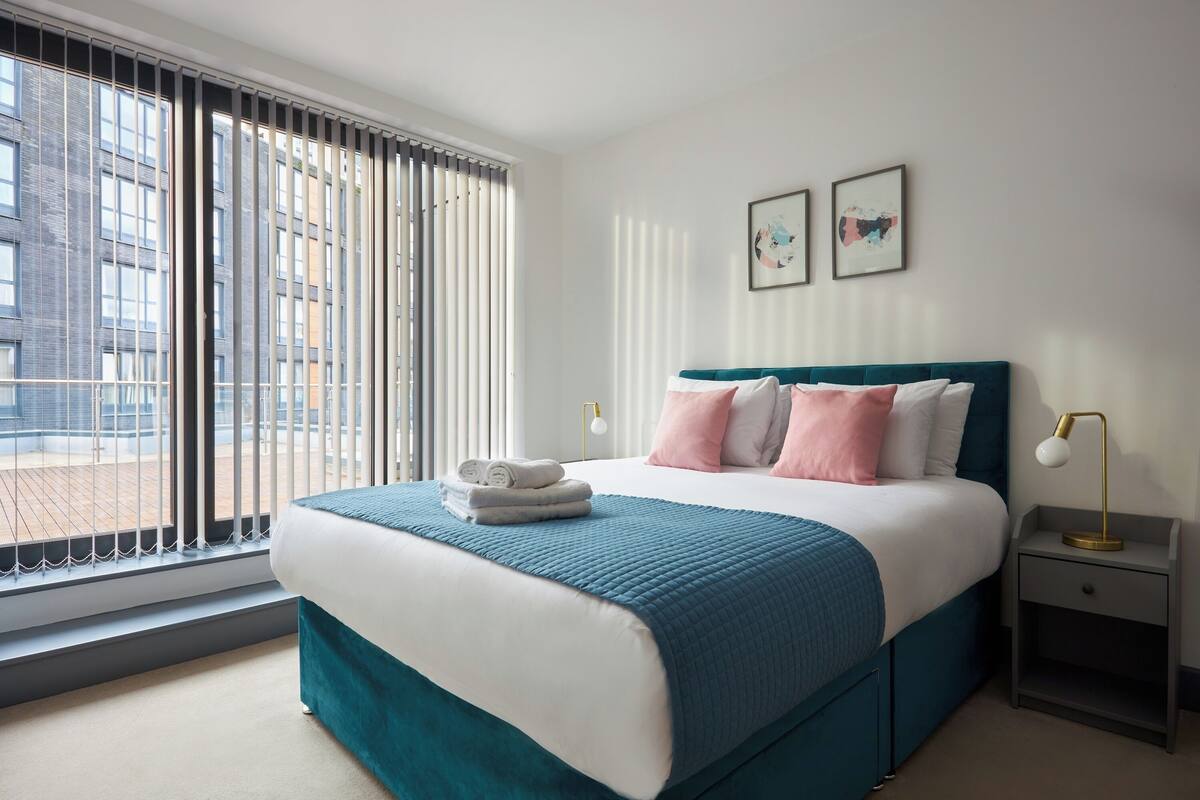 A modern hotel room with a large bed, a view through floor-to-ceiling windows, and a nightstand with a lamp.