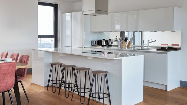 A modern kitchen with white cabinetry, a large island with stools, and a view of the city through a large window. - Canning Town by Viridian Apartments (London)