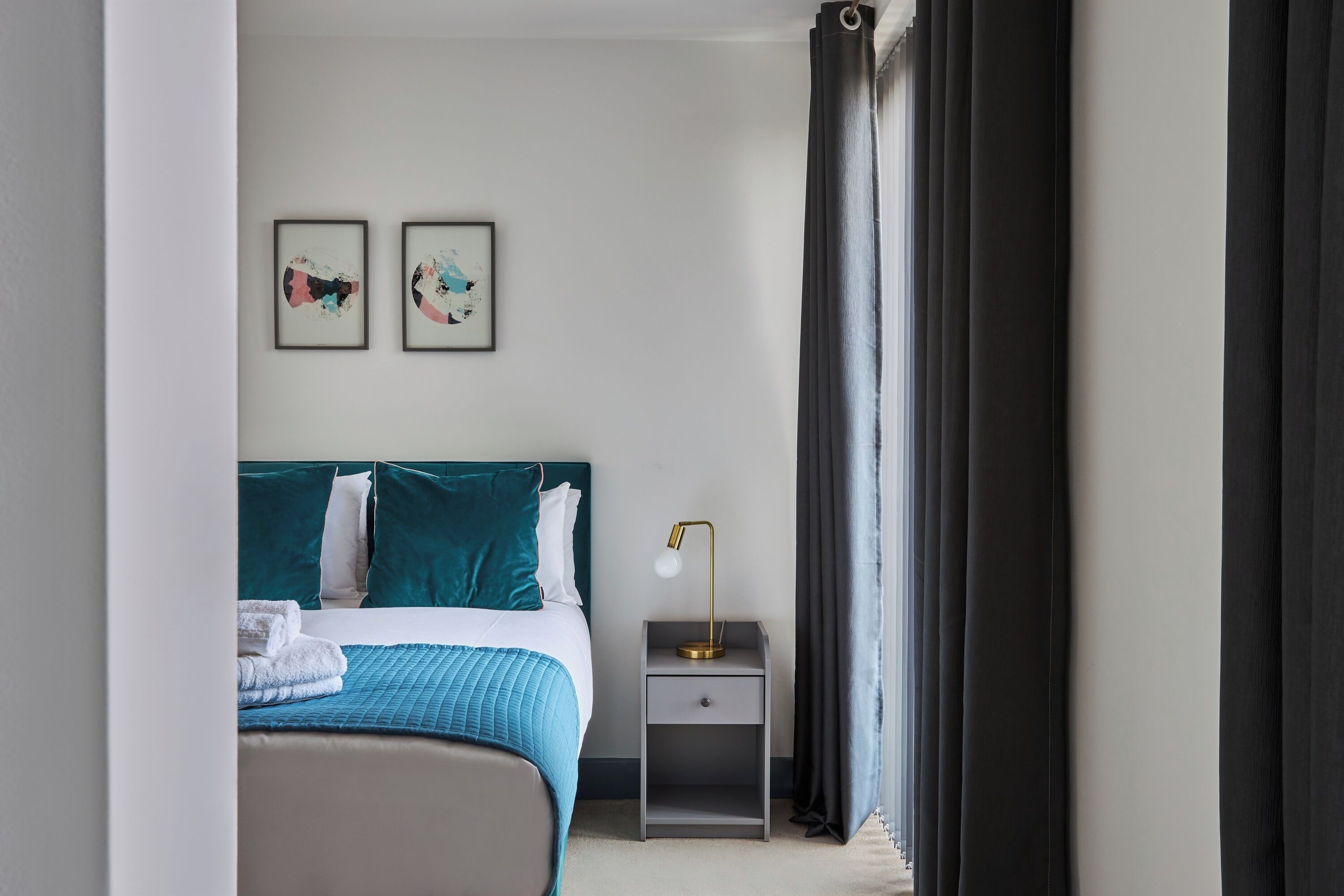 Habitación de hotel ordenada con una cama, una mesita de noche con lámpara y dos cuadros en la pared.