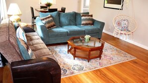 Una sala de estar con un sofá azul, un sillón marrón, una mesa de centro de cristal y una alfombra estampada.