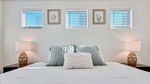 A bedroom with a bed, two bedside lamps, a window with blinds, and framed artwork on the wall.
