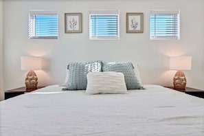 A bedroom with a bed, two bedside lamps, a window with blinds, and framed artwork on the wall.