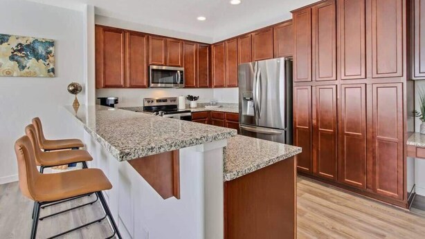 A kitchen with wooden cabinets, a granite countertop, stainless steel appliances, and a bar area with stools.