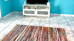 Een kamer met een blauwgroene wand, een flatscreen televisie op een witte tv-meubel, een kleurrijk gestreept vloerkleed, en decoratieve muurdecoratie van vissen en ankers.