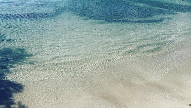 Een strand met helder turquoise water, een zandstrand en een verafgelegen landmassa begroeid met vegetatie.