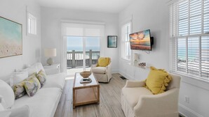 A bright living room with a large window, a sofa, a coffee table, and a TV mounted on the wall.