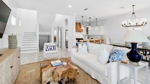 A spacious living room with a white sofa, a coffee table, and a chandelier. A staircase leads to another level.