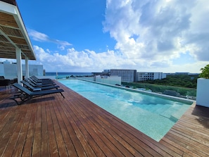 A wooden deck with a pool, lounge chairs, and a view of the ocean and buildings.
