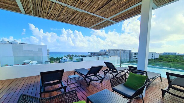 A rooftop terrace with wooden flooring, black wicker chairs with green cushions, and a view of the ocean and buildings. - Bahay By Riviera Rentals (Playa del Carmen)