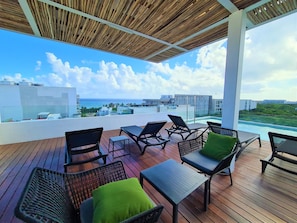 A rooftop terrace with wooden flooring, black wicker chairs with green cushions, and a view of the ocean and buildings.