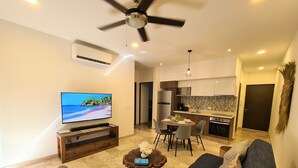 A modern living room with a flat-screen TV, a ceiling fan, and a dining area with a kitchen in the background. - Bahay By Riviera Rentals (Playa del Carmen)