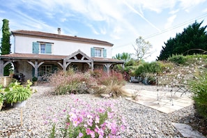 A two-story house with a tiled roof, a covered porch, and a garden with pink flowers and gravel paths. - Spacious farmhouse with private heated pool, sleeping up to 8. (Bourlens)