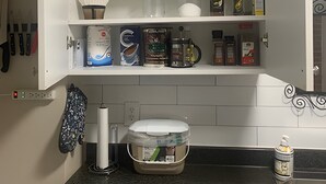 Une cuisine avec un plan de travail noir, des placards blancs et divers ustensiles, notamment une machine à café, des bocaux et des boîtes.
