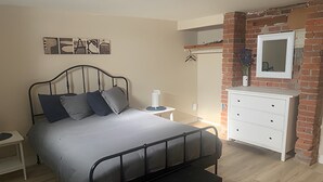 Une chambre avec un lit en métal, une literie grise, une commode blanche et un mur décoratif en briques.