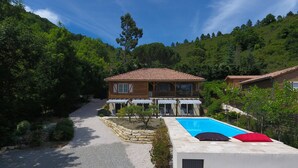 Una casa de dos pisos con piscina, rodeada por un camino de grava y abundante vegetación.