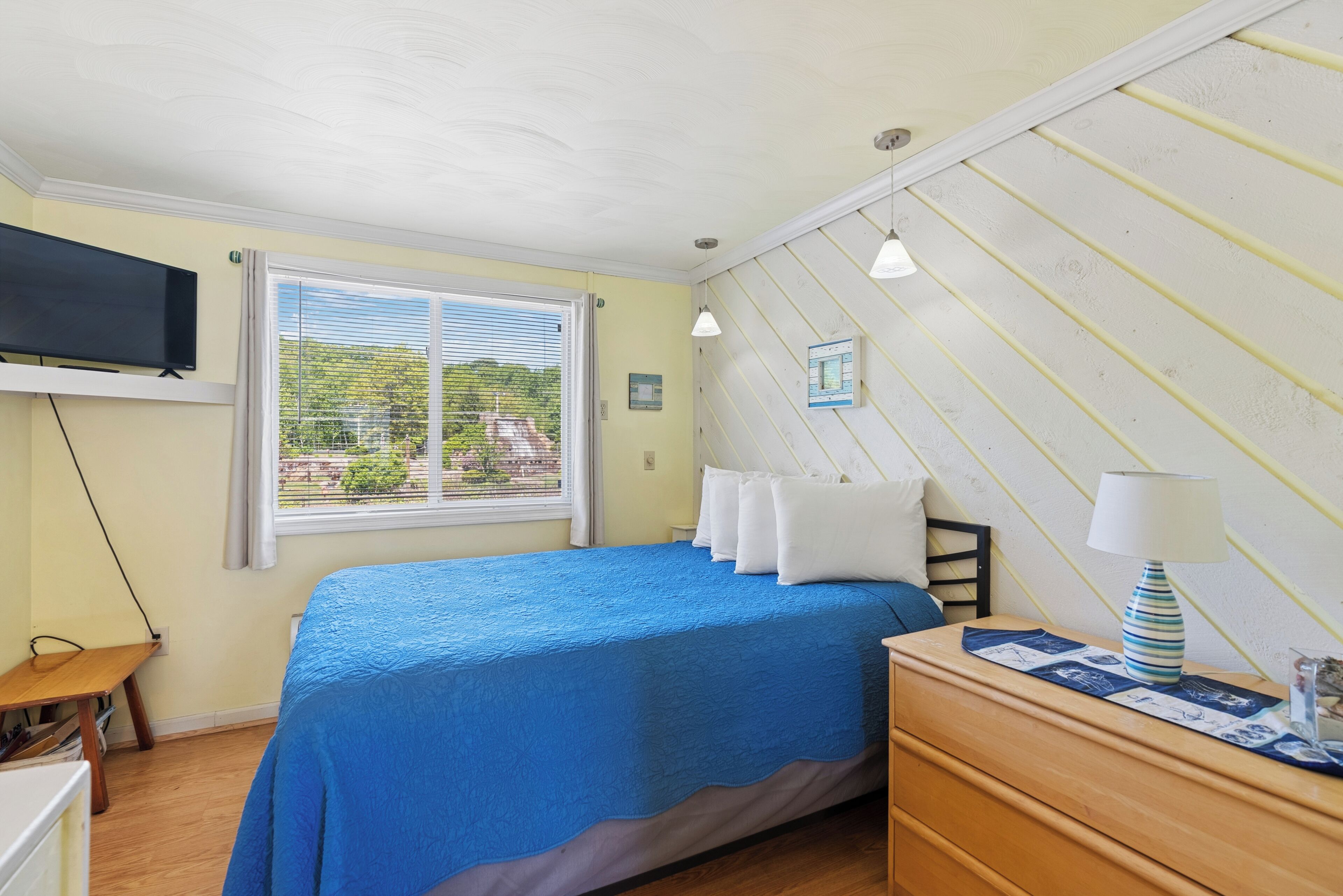 Ein Schlafzimmer mit Bett, Nachttisch mit Lampe, an der Wand montiertem Fernseher und einem Fenster mit Jalousien.