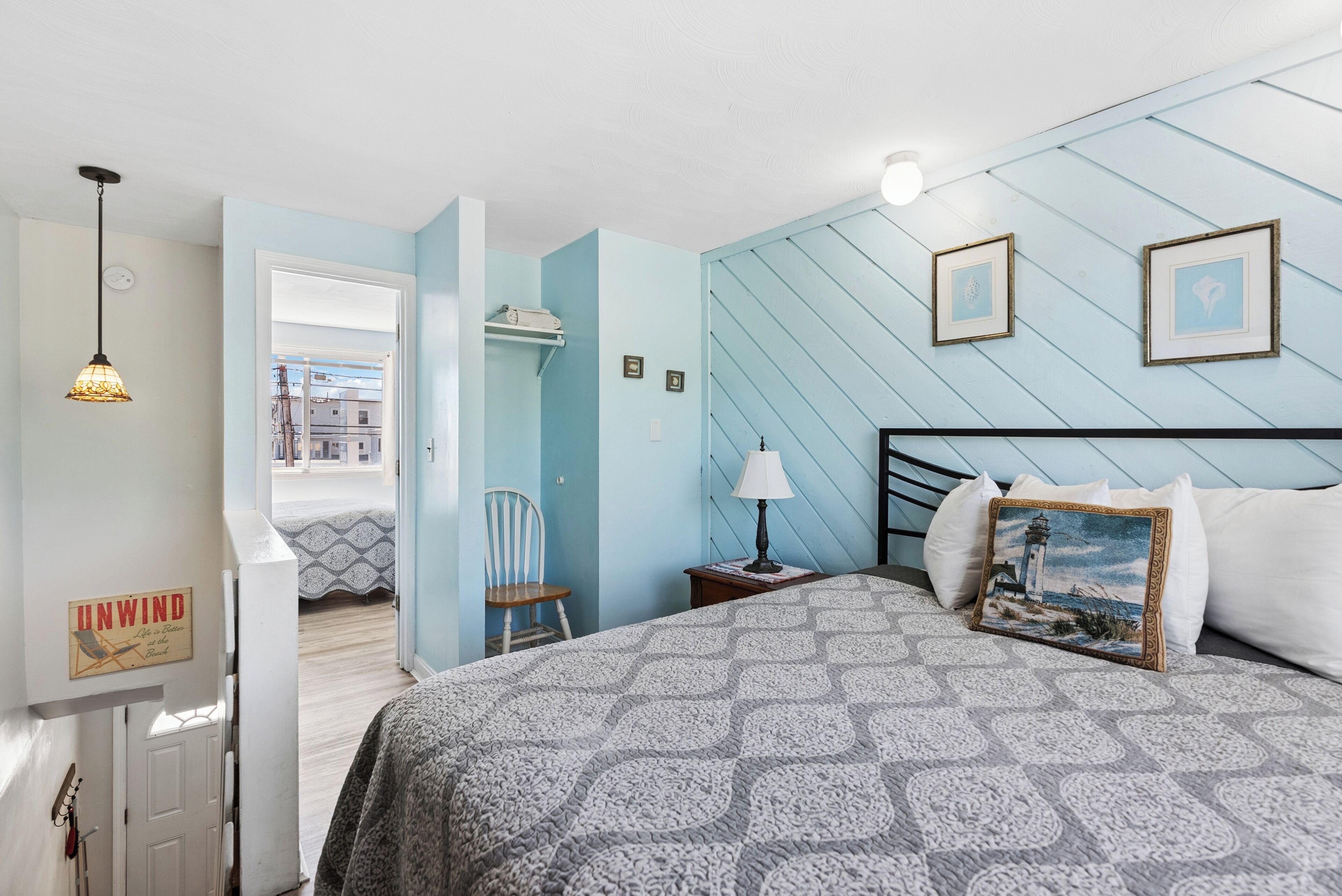 Een slaapkamer met een bedovertrek met patroon, een nachtkastje met een lamp, een ingelijst schilderij aan de muur en een raam met uitzicht op gebouwen.