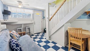 A room with a checkered floor, a wooden chair, a ceiling fan, a television, and a window with curtains. - Sands Townhouses (Old Orchard Beach)