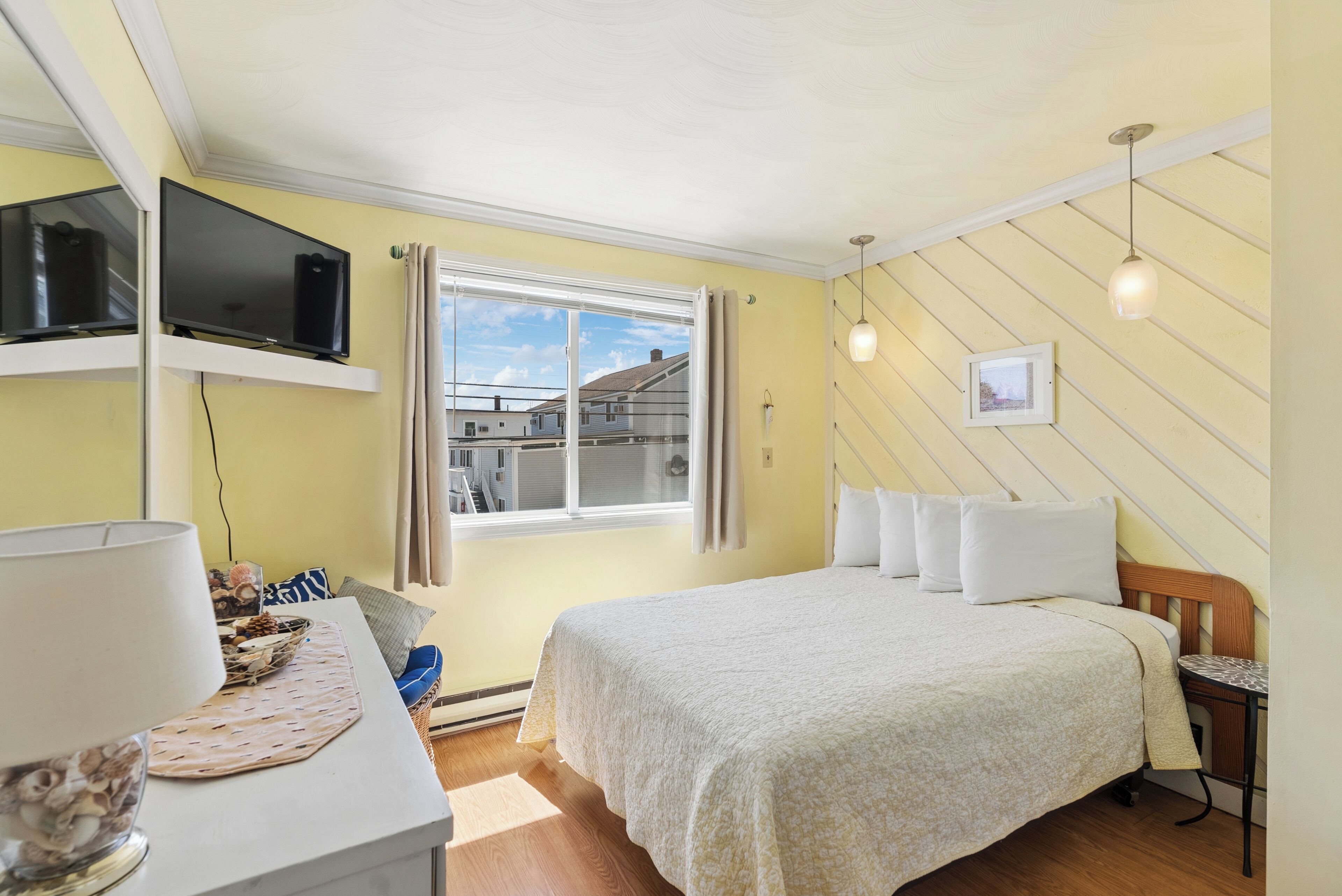 A bedroom with a bed, a TV mounted on the wall, a window with curtains, and a small table with a lamp.