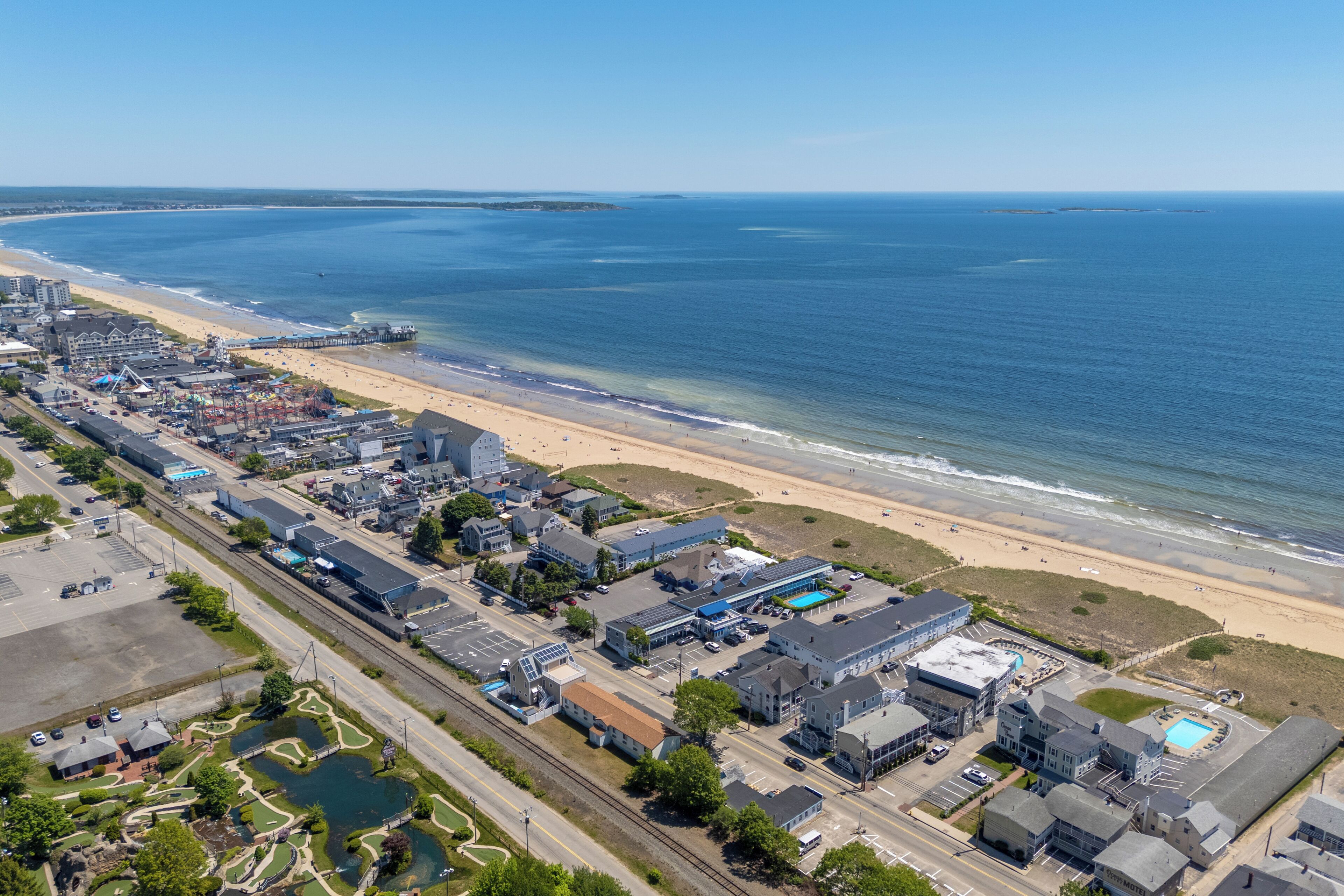 An aerial view of a coastal town with a beach, buildings, and a parking lot.