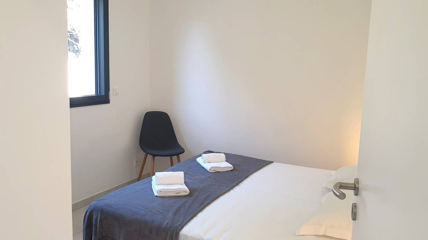 A small, clean room with a bed, two folded towels, a chair, and a window.