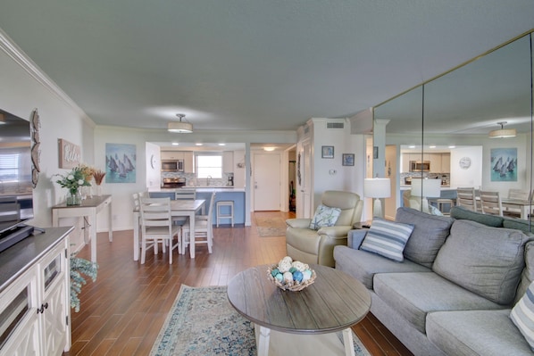 A spacious living area with a sofa, coffee table, and a dining table with chairs. A kitchen is visible in the background, and there is a large mirror reflecting the room.