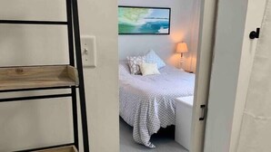 A hotel room with a wooden shelf holding towels, a bed with a patterned bedspread, a painting on the wall, and a lamp on the bedside table. - Surfs Up,  Second Row, Upper Level Unit (Folly Beach)