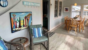 A room with a dining area, a chair with a striped pillow, a wall with a surfboard art, and a sign that says "Life is better at the beach." - Surfs Up,  Second Row, Upper Level Unit (Folly Beach)