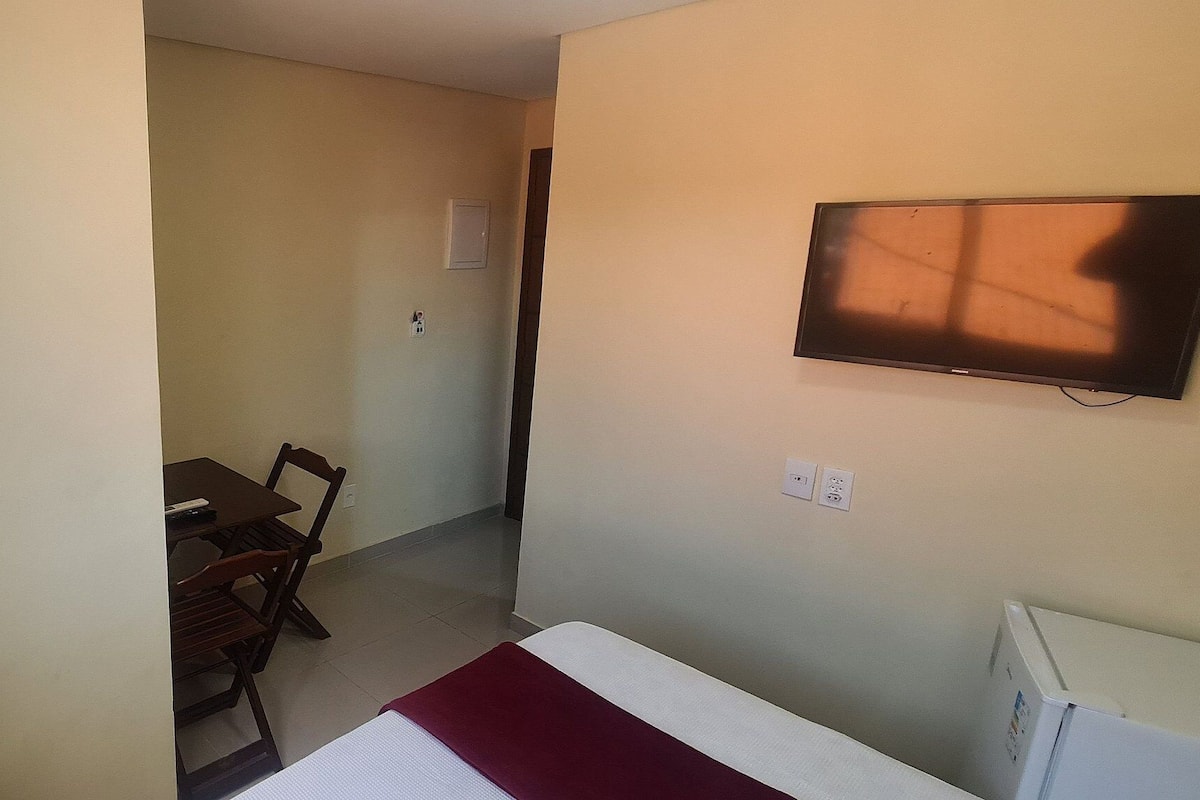 A room with a bed, a television mounted on the wall, a small table with chairs, and a window with curtains.