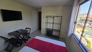 A small hotel room with a bed, a desk, a chair, a TV, and a window with a view of buildings. - Recanto Del Mare (Arraial do Cabo)