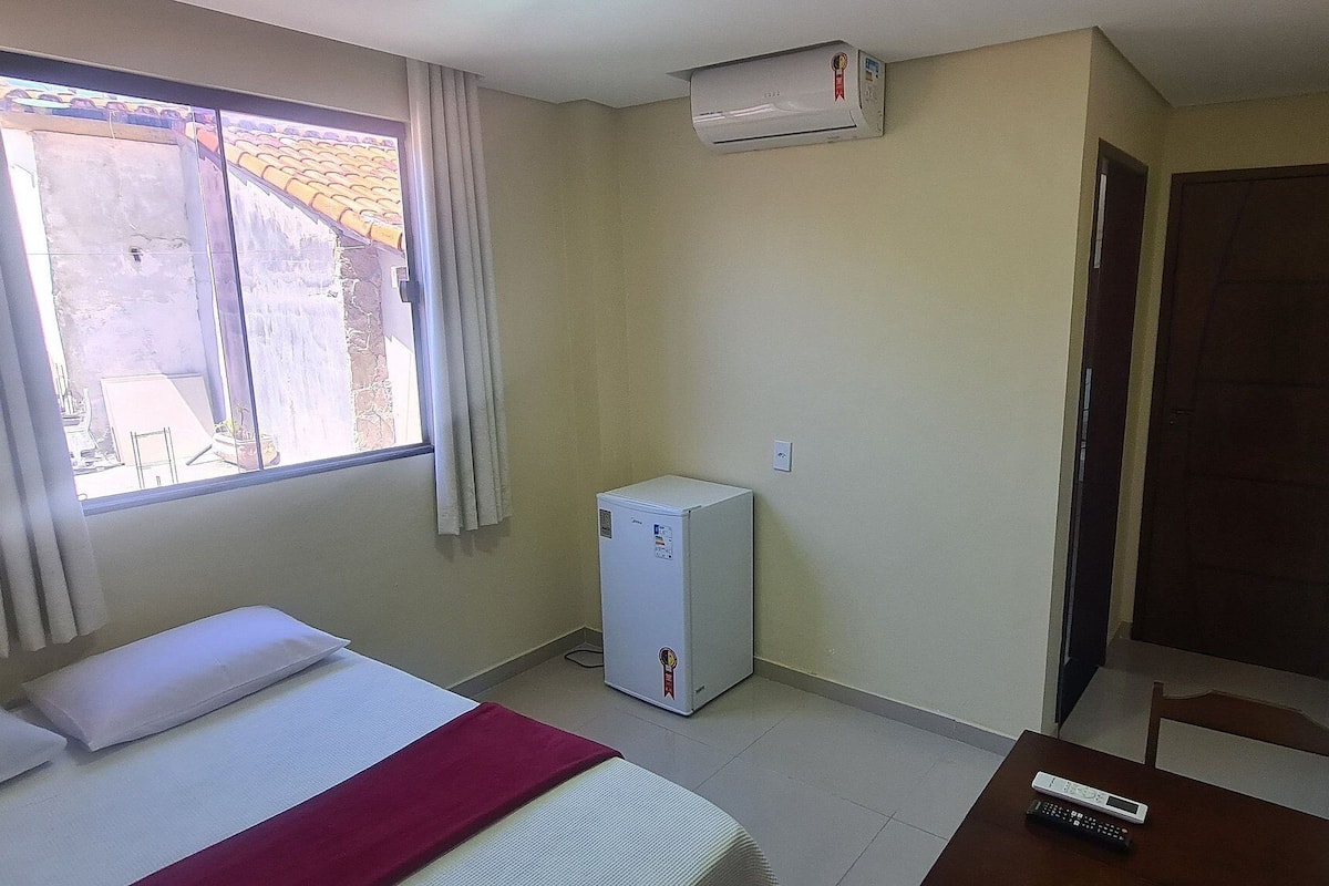 Ein Hotelzimmer mit Bett, Schreibtisch mit Fernbedienung, kleinem Kühlschrank, Fenster mit Vorhängen und einer Tür.