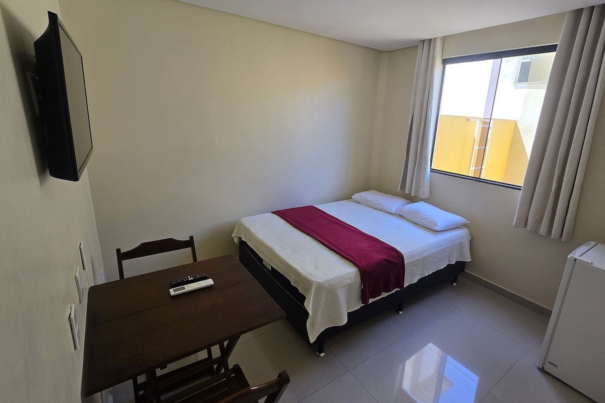A small, well-lit room with a bed, a desk, and a chair. There is a window with curtains, a flat-screen TV mounted on the wall, and a refrigerator.