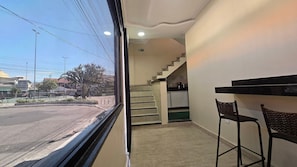 A hotel room with a view of the street, a staircase leading to the kitchen, and two bar stools.