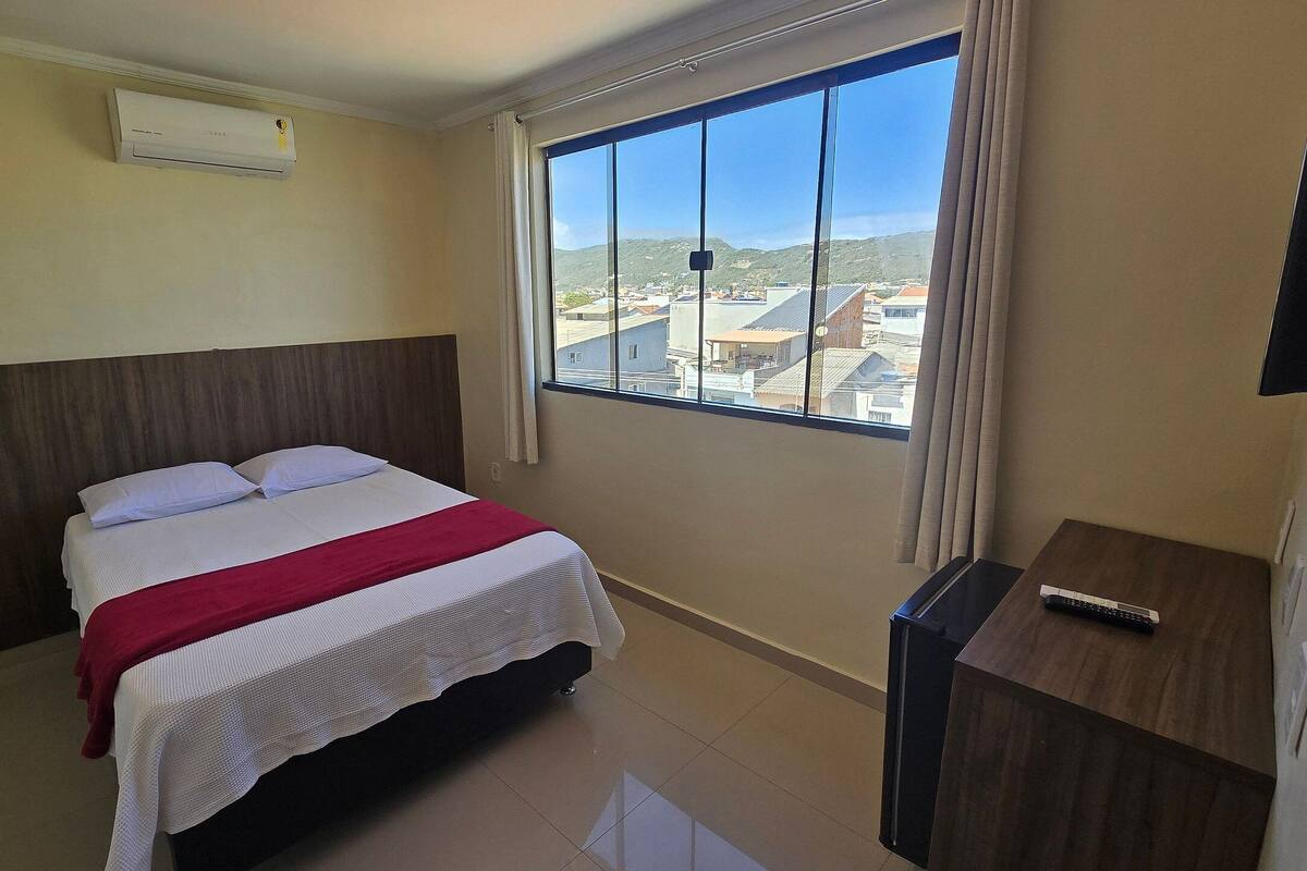 A hotel room with a bed, a TV, a window with a city view, and an air conditioning unit.
