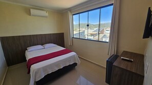 A hotel room with a bed, a TV, a window with a city view, and an air conditioning unit. - Recanto Del Mare (Arraial do Cabo)