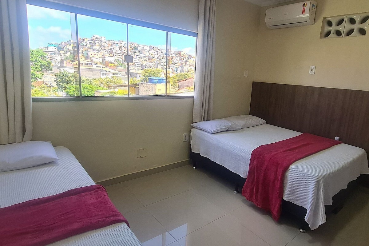 A hotel room with two beds, a window with a city view, an air conditioning unit, and a shower area.