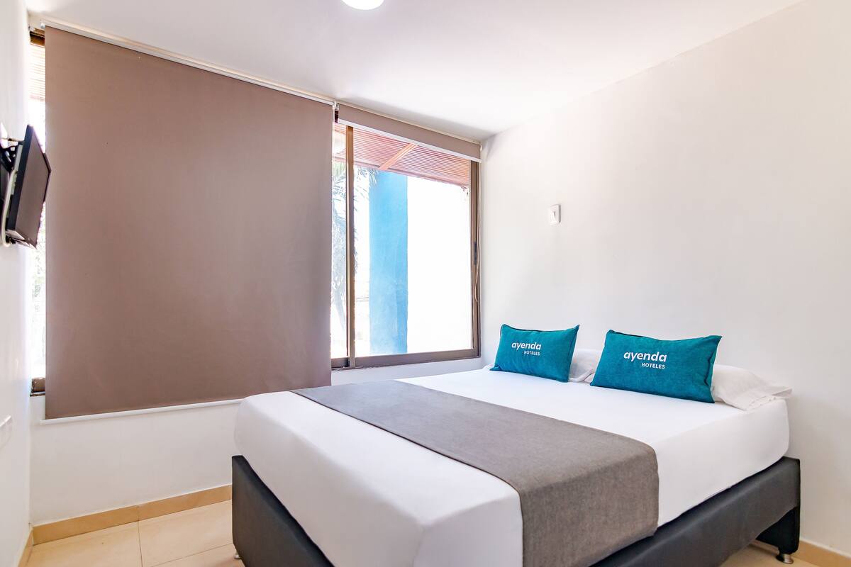 Una habitación de hotel con cama, ventana con persianas y televisor de pared.